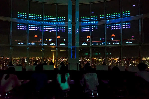 Ten players firing at Space Invaders projected across windows above Tokyo